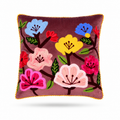 A square throw pillow with a deep plum purple velvet background, featuring colorful embroidered flowers in pink, yellow, blue, and red with green leaves, finished with a yellow pom-pom trim.