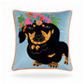 A square decorative throw pillow featuring an embroidered black dachshund wearing a colorful floral crown on a light blue background, finished with an orange pom-pom piped edge.