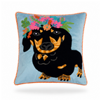 A square decorative throw pillow featuring an embroidered black dachshund wearing a colorful floral crown on a light blue background, finished with an orange pom-pom piped edge.