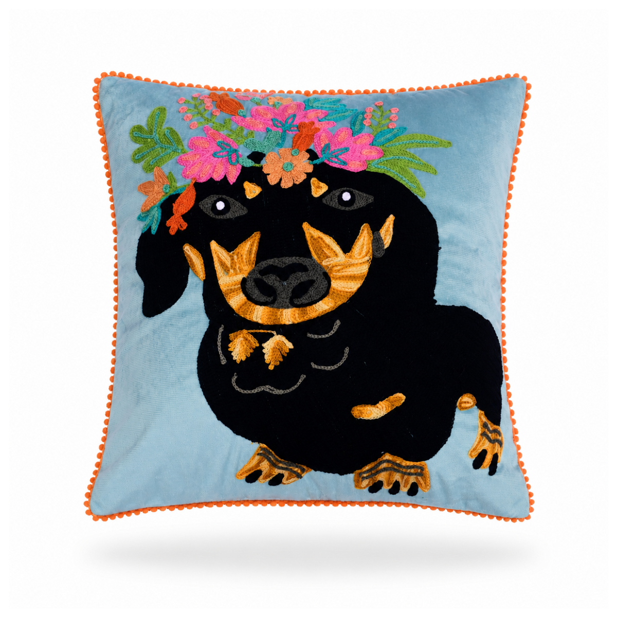 A square decorative throw pillow featuring an embroidered black dachshund wearing a colorful floral crown on a light blue background, finished with an orange pom-pom piped edge.