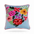 A square light blue throw pillow with a colorful embroidered floral pattern in the center and a red pom-pom trim around the edges, shown on a light blue background.