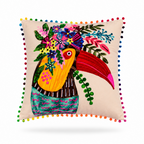 A square beige throw pillow featuring a colorful embroidered toucan holding a bouquet of flowers, finished with a border of multicolored pom-poms, shown on a white background.