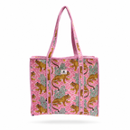A pink woven tote bag featuring an allover print of leopards and palm trees, with pink braided shoulder straps, shown against a pink white background.