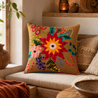 A square decorative throw pillow with a colorful embroidered floral pattern on a tan background, styled on a beige sofa in a warm, rustic room setting.