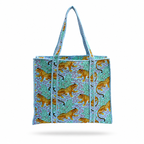 A Tote bag featuring a pattern of golden leopards on a turquoise and purple background, with light blue braided shoulder straps, shown against a plain blue purple background. 